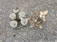 Two chandeliers placed on gravel. One has frosted glass shades and the other has metal arms with floral design elements.
