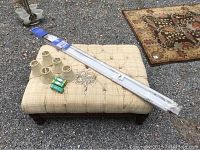 Square upholstered ottoman with tufted fabric, wooden legs, nailhead trim, placed outdoors on gravel with chandelier lamp shades, crystal pieces, and window shades on top.