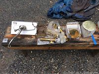 Overview of plumbing items arranged on a bench outdoors on gravel surface, showing a mix of brass and silver tone pieces including faucet fixtures, valves, and hardware.