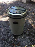 Full view of the cylindrical Soleus Air outdoor fridge, metallic body, glass lid with handle on top, standing on gravel ground outside.