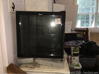 Front view of black Avanti wine cooler with glass door and metal rack inside, placed on cinder blocks in a room with various stored items.