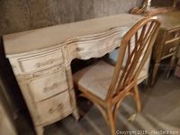 Photo of vintage distressed wooden vanity desk with matching wooden chair placed in front of it. Vanity has multiple drawers and a curved top edge.