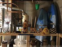 Wide view of the teapots, candlesticks, and ceramic jug arranged on a wooden shelf in an unfinished basement.