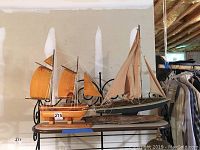Two wooden model ships on a small table against a beige wall in a basement. One ship has natural wood-colored sails and hull, the other has beige fabric sails and a darker hull on a driftwood base.
