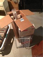 Open box showing eight rolls of Ralph Lauren wallpaper inside cardboard packaging along with a white metal rack.