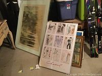 View of stacked framed pictures including large forest scene art and Chanel fashion sketch print, showing condition and styles