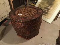 Wicker hamper closed with lid showing woven pattern, dark brown color, placed on floor next to furniture and framed picture.