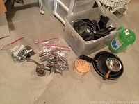 Overview of kitchen items including pots, pans, manual meat grinder, flatware in plastic bags, plastic bin with utensils, and cleaning product.