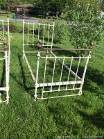 Full view of antique twin metal bed frame with white paint, brass or copper finials, side rails attached, scratches and minor dents visible.