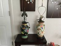 Two Asian lamps placed side by side on a dark side table against a white wall, showing detailed floral designs and double sockets on the smaller lamp.