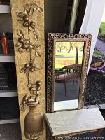 Photo showing the ornate Syrocco framed mirror leaning beside the vertical gold-toned Syrocco floral wall decor sculpture and the marble-topped stand base.