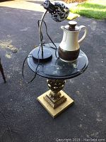 Mid century round black marble table top with gold decorative base along with a black and white gooseneck lamp and ceramic West Germany thermos teapot on the table.