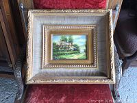 Full view of the oil painting in a large, ornate gold frame resting on wooden chair.