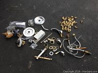 Full lot overview showing various plumbing parts on a black asphalt surface including brass knobs, chrome escutcheons, valves, and water supply lines.