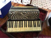 Front view of black vintage accordion showing keyboard, elaborate decorative patterns, and hard case interior.