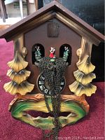 Front view of brown chalet style cuckoo clock with three figurines, carved wooden leaves and visible chain weights