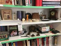 Shelf with Winchester battery shelf clock, pillar candles, brass candle holders, owl napkin holder, playing cards, VHS tapes and assorted decorative items.