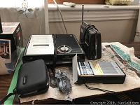 Lot of vintage electronic devices including two black transistor radios, Panasonic tape recorder with manual, TomTom GPS device, and JVC VHS player on a table