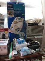 Boxed portable bath bench with attached brush, located on desk under window with air conditioner nearby. Also visible are several pill organizers, blue disposable masks, and a pair of reading glasses.