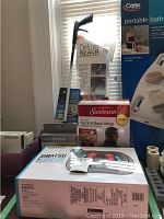 Photo showing boxed Sunbeam neck and back wrap, Carex deluxe reach assist kit packaging, portable bath seat box, and tape rolls.