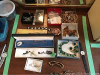 Overview of entire lot laid out showing multiple boxes and trays filled with necklaces, bracelets, pins, and other costume jewelry items including a white box displaying a guilloche cross pendant necklace and a blue box holding a green rhinestone necklace and earrings set.