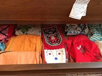 Drawer containing folded aprons in red, orange, and a black embroidered apron, as well as a note attached inside drawer