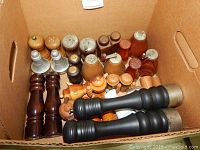 Box containing multiple pairs of salt and pepper shakers in wood, metal and aluminum, varying shapes and sizes.
