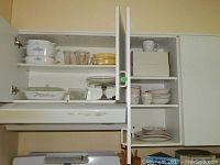 Two kitchen cabinets with assorted dishes. Left cabinet shows Pyrex casseroles, measuring cups, yellow casserole. Right cabinet shows white mugs, cups, and saucers, bowls, and stacked plates.