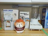 Photo showing Black & Decker JW200 automatic jar opener in original box, ceramic dog-shaped cookie jar labeled 'COOKIES', Hamilton Beach can opener box, and Hamilton Beach blender with plastic jar.