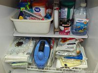 Shelf with white plastic bin holding various cleaning sprays and cleaning items, next to bottles and boxes of hand soap, vacuum bags, and Drano