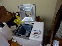 Red Star breadmaker with lid open showing the bread pan inside on a wooden surface, some background items visible.