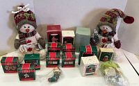 Photo of two 10-inch sitting snowman plush toys with knitted hats and scarves surrounded by boxed ornaments, showing variety and quantity in lot.
