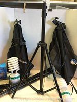 Two black Limo Studio light reflectors folded and hung against a wall with adjustable tripod stand in foreground, spiral light bulbs and partially visible blue Neewer light box.