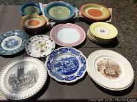 Full overview of assorted plates laid out on table showing various colors, styles, and sizes including striped concentric rings, floral, and architectural scenes.