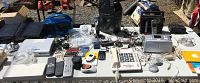 Wide table view showing multiple vintage electronic items: push button phones, calculators, remotes, cables, flashlights, photo printers, radios, USB devices, and software boxes.