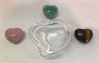 Three heart shaped stones (green, tiger's eye, light pink) and one clear glass heart shaped trinket box shown together.