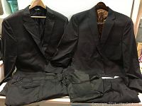 Photo of two men's dark suits laid out side by side on a flat surface, showing jackets and pants folded below.
