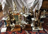 Overview of 28 vintage sports trophies arranged on a carpet and white cloth backdrop. Includes various sports figures in gold-tone atop marble or wood platforms.