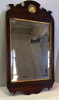 Full view of the wooden antique mirror hanging vertically on a wall, showing the dark wood frame with carved decorative crest and gold shell accent, gold inner trim, and aged mirror glass.
