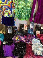 Photo showing variety of colorful ice skating dresses hanging and multiple tights, shorts, and skirts with tags scattered on surface.