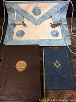 Freemason apron placed above encyclopaedia and Bible on wooden surface