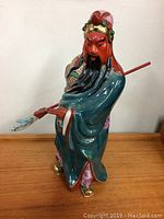 Front-left angle view of the vintage Asian figure showing a red-faced warrior in traditional attire with a pole weapon.