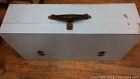 Exterior top view of the vintage blue lacquer toolbox showing its rectangular shape, blue color, brown handle, and metal hinges and clasp on a wooden floor.