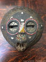 Front view of round wooden mask showing bead decorations around eyes, nose and mouth with inset metal pieces.