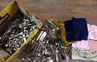 Large box filled with mixed silver plate forks, spoons, knives and fabric holders laid beside it on wooden surface.