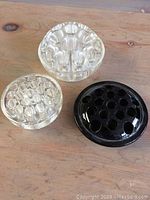 Three glass flower frogs on wooden surface: two clear and one black. The clear ones include one flat circular with grid holes and one rounded frog-shaped. The black one is flat circular with holes.
