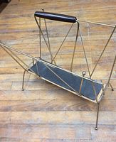 Side angle view of vintage mid century modern magazine rack with brass/gold tone metal frame, wooden handle, and black panel base on wooden floor.