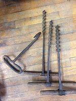Photo showing one vintage metal hook and three large spiral drill bits laid flat on a wood surface.