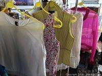 Photo showing white, floral, yellow striped, white lace sleeveless tops, and pink sheer top hanging on yellow hangers.