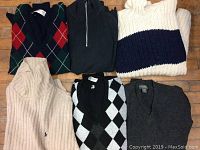 Photo showing six men's sweaters of various colors and styles, laid out on wooden floor.